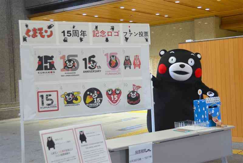 15周年記念ロゴに関するファン投票を呼びかけるくまモン。ボードに掲げられた10のロゴ案も紹介した＝3日、熊本県庁