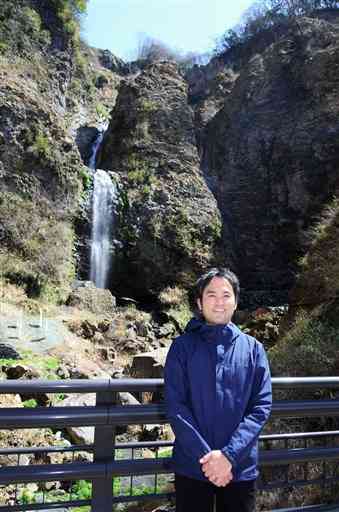 垂玉温泉の泉源を生んだとされる金龍の滝（後方）の前で笑顔をみせる山口雄也さん＝３月２４日、南阿蘇村