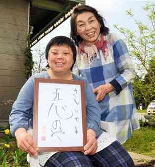 自分で書いた「五心のやさしいです」の文字を手に、ほほ笑む和田朋子さん。後ろは母親の緑さん＝菊池市