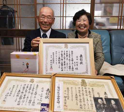 熊日金婚夫婦の表彰状を手に笑顔の愛甲利孝さん（左）と文子さん。利孝さんの祖父母、両親の分と合わせ、３代の表彰状がそろった＝あさぎり町