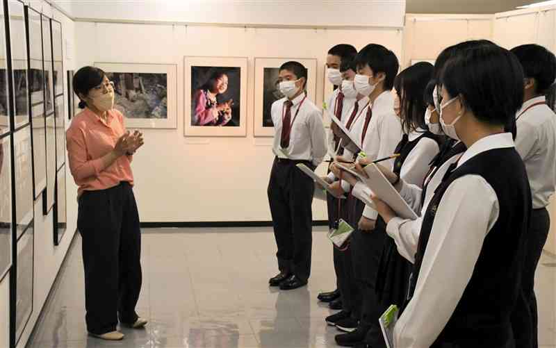 「9人の写真家が見た水俣」展の会場を訪れ、作品の時代背景について説明を受ける熊本中央高の生徒たち＝18日、熊本市中央区