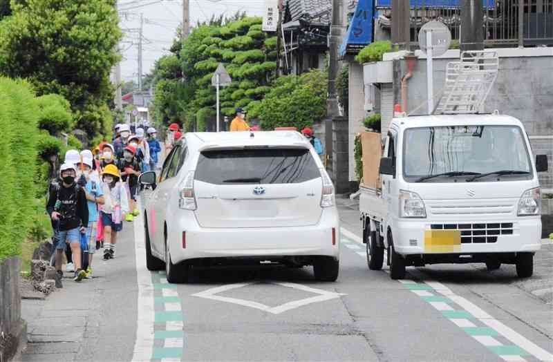 熊本市南区城南町の市道で、対面通行する2台の車の横を歩く児童ら＝５月20日午前7時40分ごろ（写真を一部加工しています）