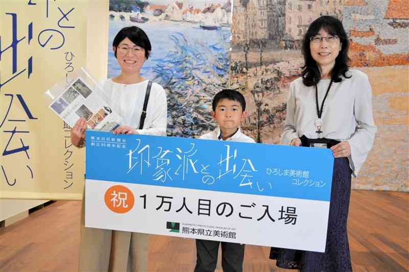 「印象派との出会い」展で1万人目となり、早田章子館長（右端）から図録を受け取った家族連れ＝26日、熊本市中央区