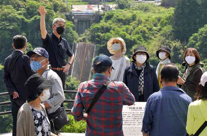 熊日東京支社の小多崇編集部長（左上）から熊本地震の被災状況などの説明を受けるバスツアーの参加者ら。奥は崩落した阿蘇大橋＝29日午前、南阿蘇村立野（後藤仁孝）