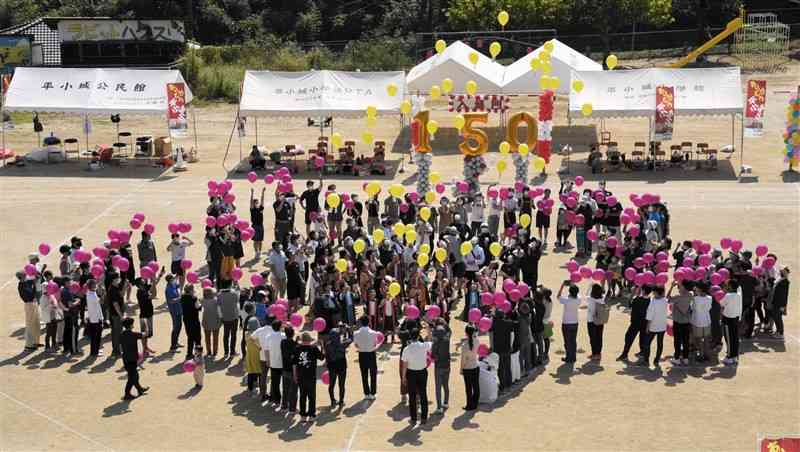 校章の桜の花をかたどって並ぶ地元住民と黄色の風船を飛ばす平小城小の児童＝山鹿市