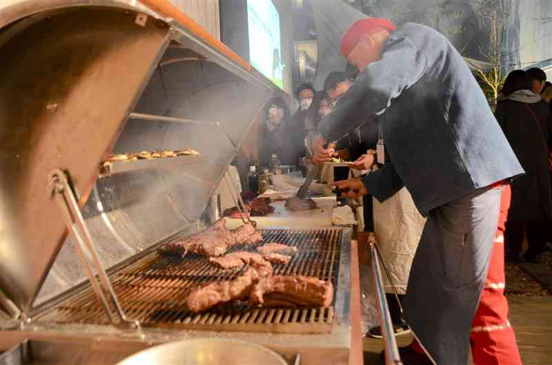 あか牛が焼き上がるのを待つ参加者ら（奥）＝熊本市中央区