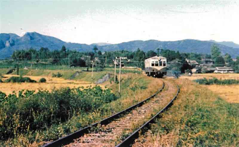 1959年、現在の山鹿市鹿本町藤井付近の田園地帯を走る鹿本鉄道のディーゼル車両。左奥は山鹿名所の不動岩（西村マサ子さん提供）