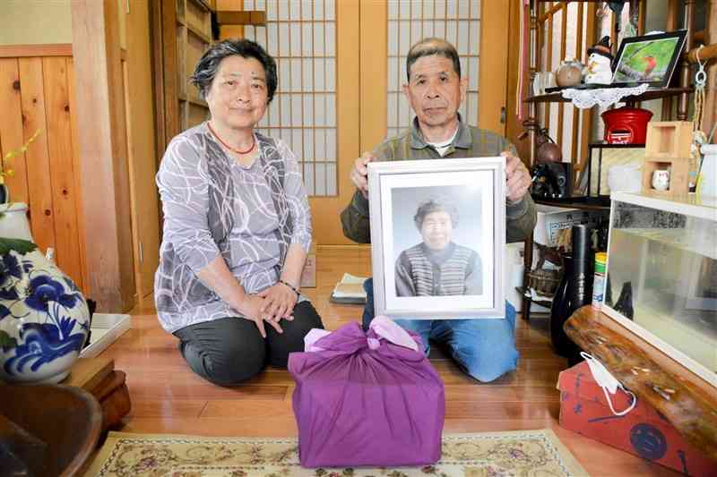 城幸惠さんの写真を持つおいの文博さんと妻の裕子さん。手前の風呂敷包みは、幸惠さんの記事が掲載された新聞記事＝17日、芦北町