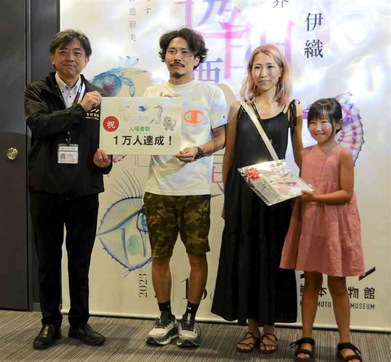 「新世界『透明標本』展」の来場1万人目となり、坂本康祐館長（左）からグッズを贈られた女子児童（右）ら＝熊本市中央区