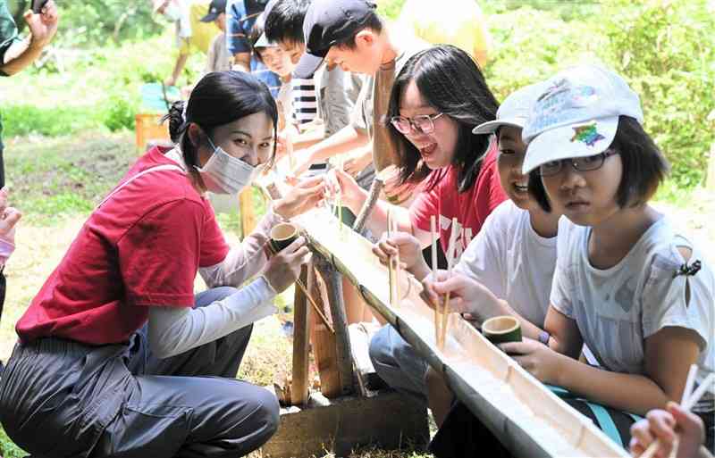 小学生らとそうめん流しを楽しむ台湾・淡江大のインターン生2人（赤いTシャツ）＝阿蘇市