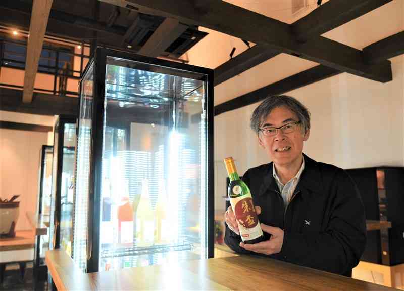 千代の園酒造が開業する立ち飲み店。ショーケースの冷蔵庫前で大吟醸「朱盃」のボトルを持つ本田雅晴社長＝22日、山鹿市