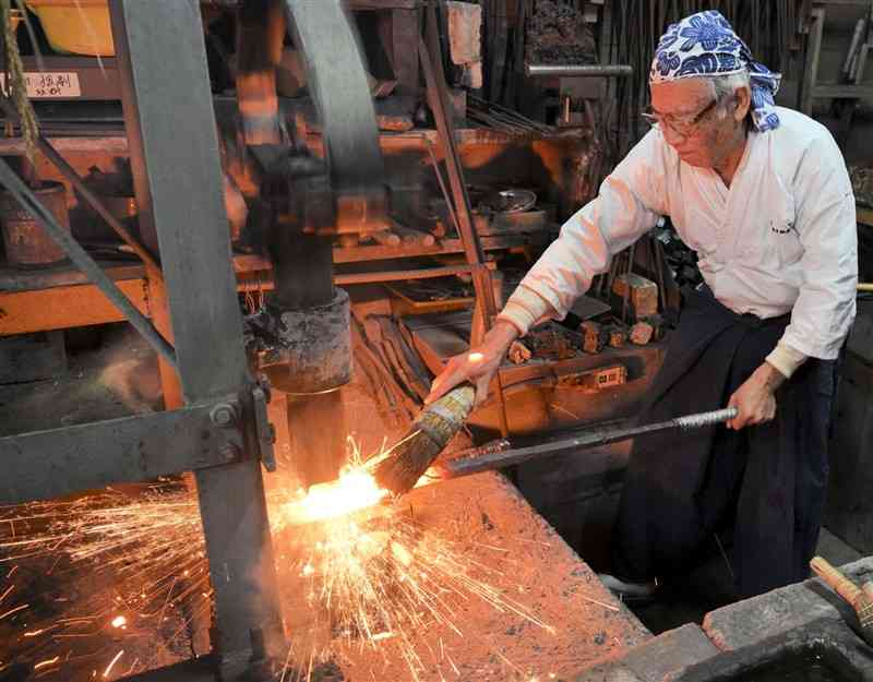 赤く熱した玉鋼を機械式のハンマーで鍛錬する松永源六郎さん＝3日、荒尾市