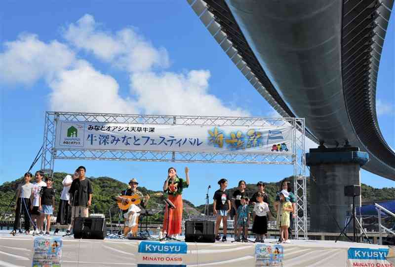 牛深ハイヤ大橋の真下のステージで、歌謡曲「にじ」を歌うMAYUMEさん（中央）と子どもたち＝21日、天草市