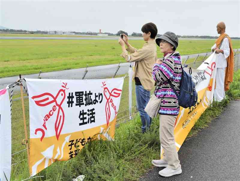 日米共同統合演習の中止を求める横断幕を掲げ、滑走路を見つめる和田知子さん（手前）ら＝23日、熊本空港