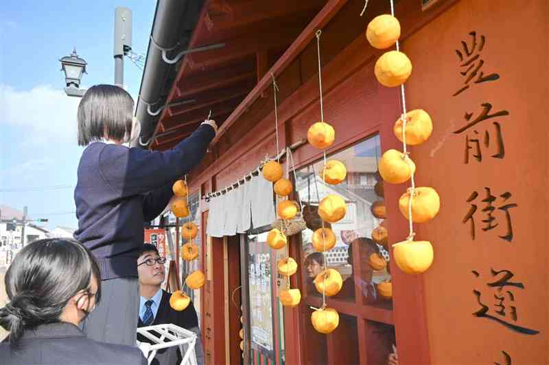 豊前街道沿いのカフェの軒先に、干し柿をつるす城北高の生徒たち＝3日、山鹿市