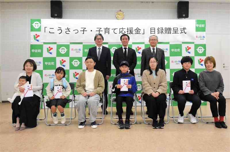 甲斐高士町長（後列中央）から応援金の目録を受け取った受給対象の親子ら（前列）＝7日、甲佐町