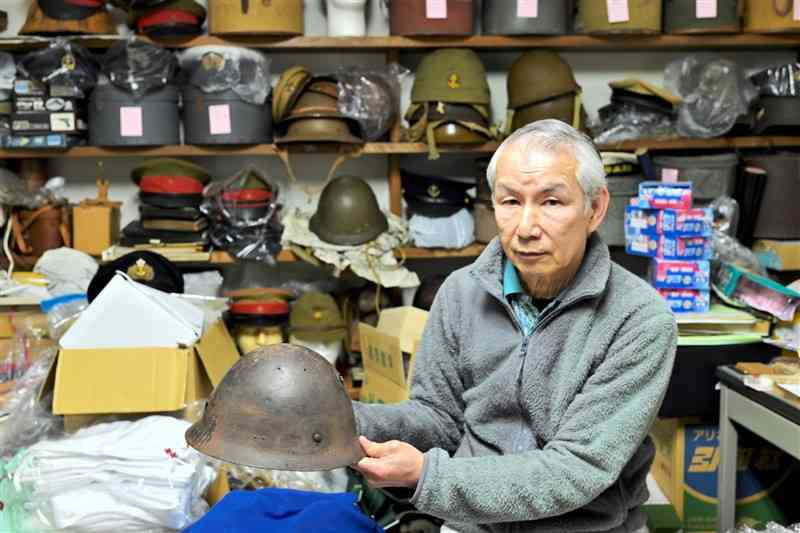 太平洋戦争中の軍装品や日用品の収集を半世紀近く続ける松山強さん。手にする鉄かぶとは銃弾が額から後頭部に向けて貫通した痕が残る＝1月27日、荒尾市