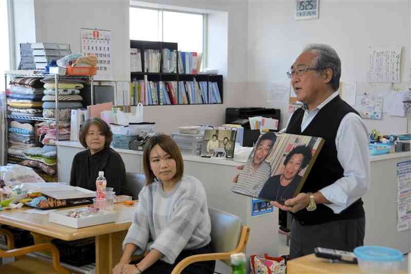 川本輝夫さんと妻のミヤ子さんの写真を手に、あいさつする長男の愛一郎さん（右）＝16日、水俣市